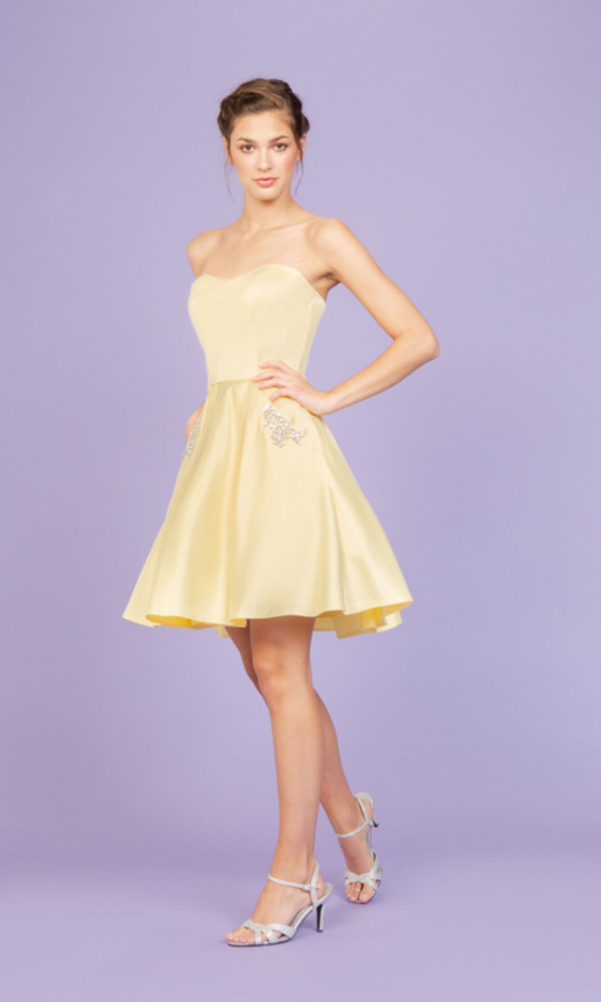 A woman poses against a light purple background in the Strapless Short Wedding-Guest Dress: Eureka 6622, a pale yellow, knee-length cocktail dress with a strapless neckline, paired with silver heels.